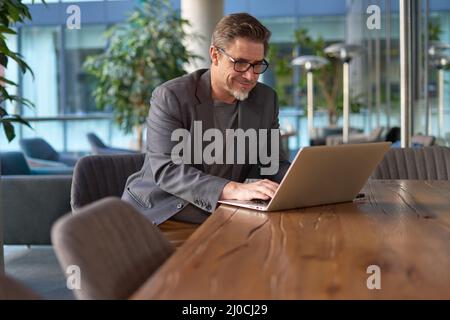 Glücklicher Geschäftsmann mittleren Alters, der im Büro einen Laptop benutzt. Ein älterer, zwangloser Mann, der online auf der Terrasse des Cafés arbeitet und lächelt. Stockfoto