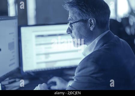 Geschäftsmann, der am Schreibtisch mit einem Computer arbeitet, denkt nach. Porträt eines Mannes mit mittlerem Erwachsenen in Hemd und Jacke, lächelnd. Bärtig, Brille. Stockfoto