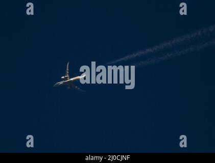 Ein kommerzielles Verkehrsflugzeug mit Kondensstreifen in großer Höhe fliegt bei Sonnenuntergang durch einen tiefblauen Himmel. Stockfoto