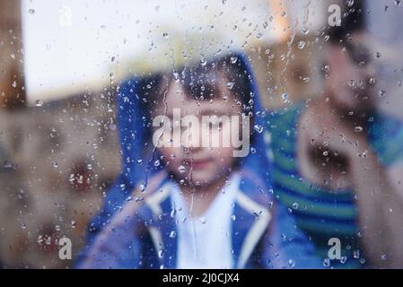 Regnerische Tage ohne viel zu tun Ein kleiner Junge schaut nach unten hinter einem durch Regen gestreift Fenster, während seine Mutter im Hintergrund sitzt. Stockfoto