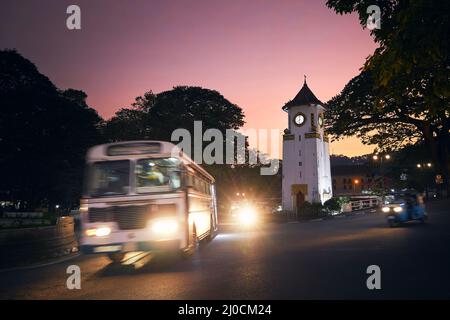 Bus und Tuk Tuk in unscharfer Bewegung an der Kreuzung bei schönem Sonnenuntergang. Verkehr rund um die Uhr Turm auf der Straße in Kandy, Sri Lanka. Stockfoto