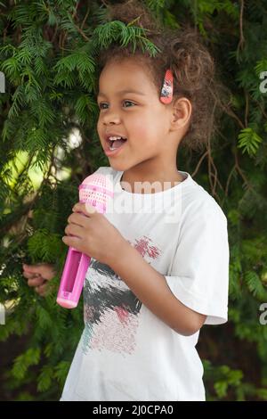 Nette kleine Sängerin Mädchen singen auf dem Mikrofon im Stadtpark von grüner Vegetation umgeben