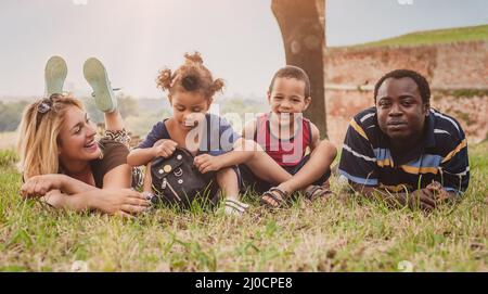 Outdoor-Porträt einer glücklichen internationalen Familie, die Spaß hat und auf einem grünen Gras im Park liegt Stockfoto