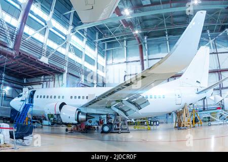 Weißes Passagierflugzeug im Hangar. Flugzeuge, die in Wartung sind. Überprüfung der mechanischen Systeme für den Flugbetrieb Stockfoto