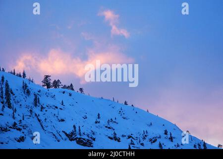 Das Licht des Sonnenuntergangs trifft auf eine wispige Wolke über einem schneebedeckten Berg Stockfoto