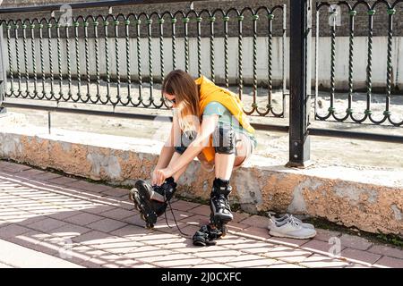 Junge Frau im gelben Hoodie, die Schutzausrüstung anlegt, bevor sie am Ufer Rollschuhlaufen kann aktiver Lebensstil, Outdoor-Aktivitäten, Rollschuhlaufen Stockfoto