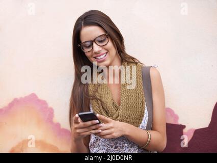 Shes immer verbunden. Eine wunderschöne junge Frau, die ihr Handy an einer mit lebendigen Farben bemalten Wand anhat. Stockfoto