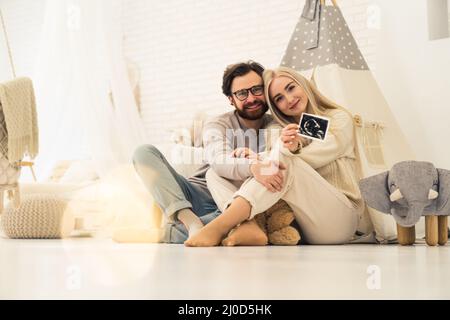 Schönes kaukasisches Paar, das zusammen sitzt und ein Ultraschallfoto ihres ungeborenen Babys zeigt. Hochwertige Fotos Stockfoto