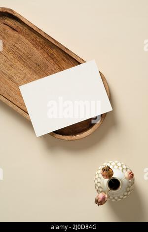 Unbeschriftete Papierkarte auf Holztablett mit Vase mit getrockneten Blumen auf beigefarbenem Hintergrund. Flach liegend, Draufsicht, Kopierbereich. Stockfoto