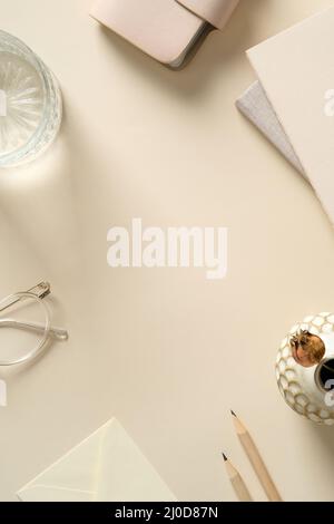 Ästhetischer femininer Arbeitsplatz mit Papierheften, Glas Wasser, Brillen, Umschlag, Bleistiften auf beigem Hintergrund. Stockfoto