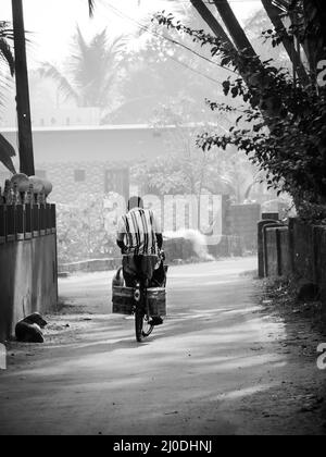 Malvan, Indien - 20. Dezember 2021 : Indischer männlicher Dorfbewohner, der auf einem Fahrrad in einer kleinen Gasse des Dorfes Konkan, umgeben von Kokosnussbaum, fährt Stockfoto