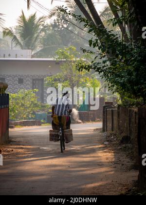 Malvan, Indien - 20. Dezember 2021 : Indischer männlicher Dorfbewohner, der auf einem Fahrrad in einer kleinen Gasse des Dorfes Konkan, umgeben von Kokosnussbaum, fährt Stockfoto