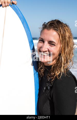 Kaukasische Surferin mit einem Frauenportrait am Strand, das Surfbrett in einem Neoprenanzug hält Stockfoto