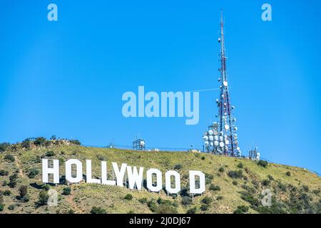 Das berühmte weiße Hollywood-Schild in den Hügeln von Los Angeles, Kalifornien, eingerahmt von einem massiven Zellenturm und einem strahlend blauen Himmel.
