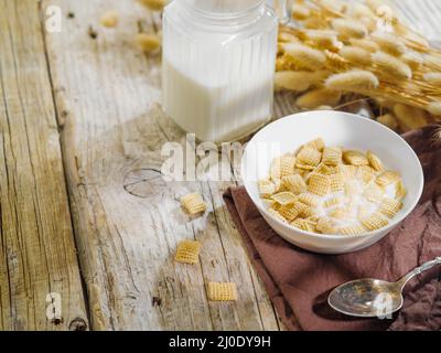 Eine Schüssel mit Müsli-Pads gefüllt mit Milch, ein Krug mit Milch, ein paar Müsli auf einem hölzernen Hintergrund. Solarbeleuchtung. Schnelles Frühstück, Cerealien, Bio-Qualität Stockfoto