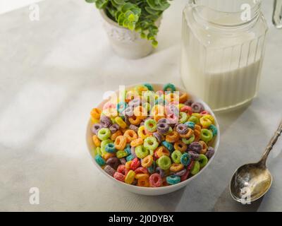 Ein schnelles Cerealien-Frühstück - fruchtscharbene Ringe in einer Schüssel, ein Löffel, ein Krug Milch und Dekor - eine Innenblume. Isoliert auf weißem Hintergrund. Niedriger Winkel Stockfoto