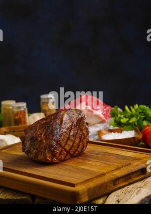 Appetitanregende Schinken im Vordergrund auf einem Holzbrett, im Hintergrund - rohes Fleisch, Kräuter, Gemüse, Gewürze. Dunkelblauer Hintergrund. Festliches Gericht. Resta Stockfoto