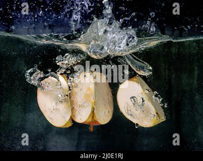 Auf dunklem Grund fallen in Scheiben geschnittene Äpfel ins Wasser. Unterwasseransicht. Kochen, vegetarisch, Kochen zu Hause Konzept Stockfoto