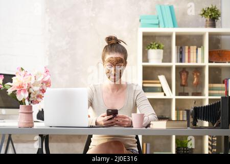 Frau kosmetische Gesichtshaut Pflege. Frau in der Haut Gummi Peeling Gesicht Schönheitsmaske sitzt am Schreibtisch mit Telefon und Laptop im Home Office. Stockfoto