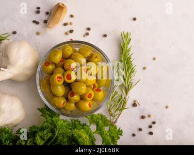 Grüne Oliven in einer Glasschüssel, Kräuter, Knoblauch auf weißem Hintergrund mit kleinen schwarzen Pfefferkörnern. Low-Angle-Ansicht. Kochen, Essen, Snacks, Vitamine, Antio Stockfoto