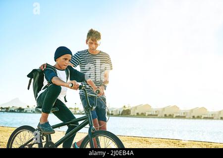 Seinem Bruder beibringen, wie man Fahrrad fährt. Ein kleiner Junge, der seinem jüngeren Bruder beibringt, wie er mit dem Fahrrad an einer Lagune entlang fährt. Stockfoto