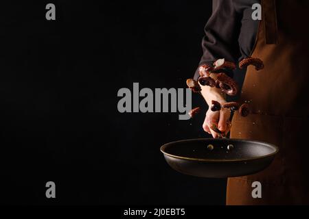 Ein professioneller Koch in dunkler Uniform kocht Krakenstücke in einer Pfanne auf einem gefrorenen Flug. Isoliert auf schwarzem Hintergrund. Restaurant, Hotel, Bankett, Koch Stockfoto