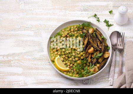 Geschmorte Kichererbsen in grüner Sauce mit gebackener Aubergine, Petersilie und Zitrone auf weißem Holzhintergrund, rustikaler Stil, Draufsicht. Leckeres gesundes Zuhause Stockfoto