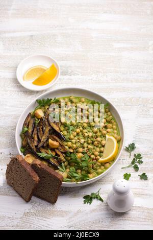Geschmorte Kichererbsen in grüner Sauce mit gebackener Aubergine, Petersilie und Zitrone auf weißem Holzhintergrund, rustikaler Stil, Draufsicht. Leckeres gesundes Zuhause Stockfoto