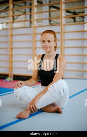 Porträt einer fitgen Frau, die auf einer Sicherheitsmatte in türkischer Sitzposition sitzt. Turnhalle-Hintergrund. Lächelnd zur Kamera Stockfoto