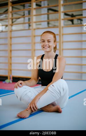 Porträt Sportliche Rothaarige junge Frau, die auf dem Boden sitzt und lächelt zur Kamera. Entspannung nach dem Training. Stockfoto