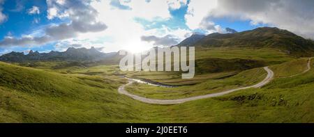 Panoramabild eines von hohen Bergen umgebenen Tals in den französischen Pyrenäen mit Wiesen, einer kurvenreichen Straße und einem Bach, bewölktem Himmel und Sonne Stockfoto