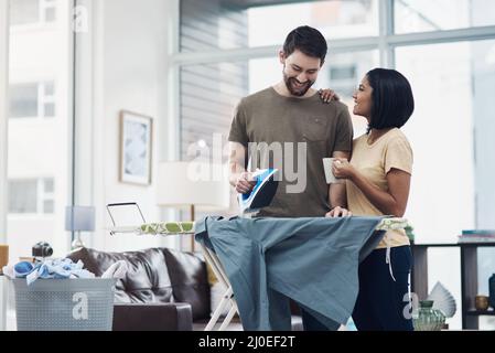 Die Aufteilung der Aufgaben ist der einfachste Weg zur häuslichen Glückseligkeit. Ein glückliches junges Paar, das zu Hause frisch gewaschene Wäsche bügelt hat. Stockfoto