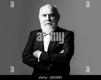Alter Mann mit gekreuzten Armen. Grauhaarige Senior Business Mann trägt eleganten Anzug und Krawatte über grauen Hintergrund, lächelnd zuversichtlich. Stockfoto