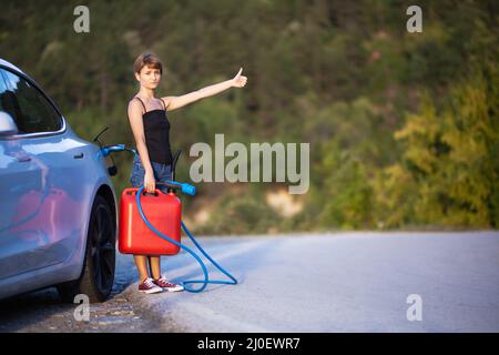 Mädchen, das neben einem Elektroauto steht, hält ein Ladekabel und einen Gassolinenkanister, während es mit dem Anhalter unterwegs ist. Stockfoto