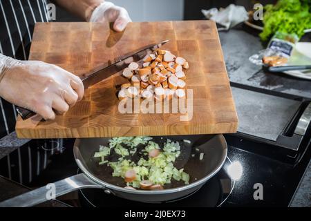 Der Koch überträgt die gehackten Zwiebeln und Würstchen in eine beheizte Bratpfanne Stockfoto