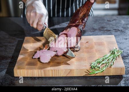 Ein Koch schneidet ein geräuchertes Lammbein in Scheiben auf einem hölzernen Schneidebrett Stockfoto