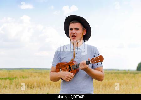 Fröhlicher Mann. Junger schöner Kaukasischer, der draußen Ukulele spielt. Schöner Hipster Kerl spielt Ukulele Gitarre auf Natur Hintergrund. Männchen in einem Cowboy Stockfoto