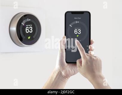 Eine Person, die eine Smartphone-Anwendung nutzt, um Energie zu sparen, mit einem drahtlosen intelligenten Thermostat auf weißem Hintergrund. Stockfoto