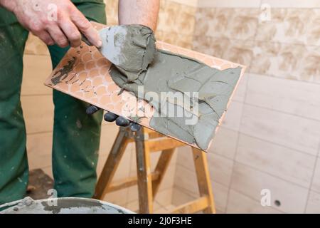 Ein Bauarbeiter trägt Zementkleber auf eine Keramikfliesen auf und verteilt ihn gründlich über die gesamte Oberfläche. Stockfoto