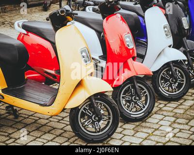 Elektromotorräder in verschiedenen Farben stehen in einer Reihe auf einer Steinpflasterung. Stockfoto