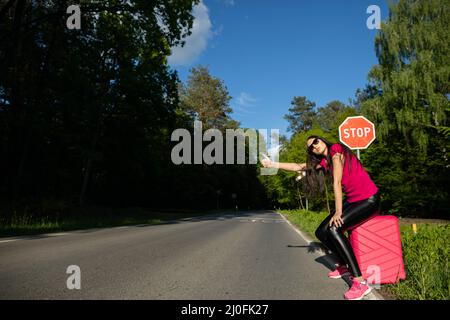 Ein junger Erwachsener steht mit einem rosafarbenen Koffer am Straßenrand und versucht, die anderen Autos anzuhalten, um eine freie Fahrt zum Hol zu bekommen Stockfoto