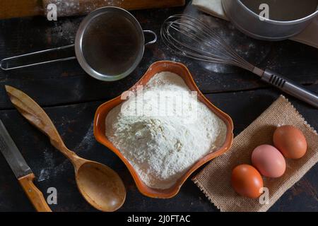 Küchengeräte und Lebensmittel auf dem alten Desktop. Weizenmehl in einer Keramikschale und Hühnereier für die Stockfoto