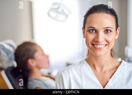 So froh, dass ich mich für die Zahnmedizin entschieden habe. Eine Zahnärztin, die in ihrem Büro steht, mit ihrem Kinderpatienten im Hintergrund. Stockfoto