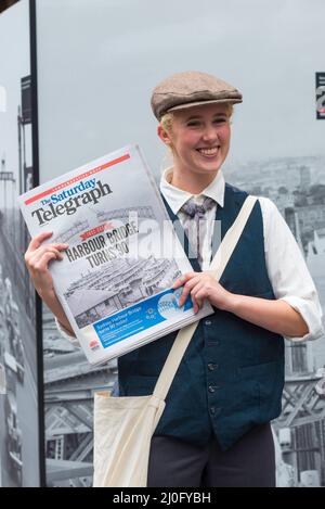 19.. März 2022, Sydney Australien: Eine Frau, die als Papierjunge der 30er Jahre gekleidet ist, gibt am Tag der 90.. Geburtstagsfeier der Eröffnung der Sydney Harbour Bridge die Zeitung Saturday Telegraph (Murdoch Press) frei. Der Bau der Brücke dauerte 8 Jahre und wurde 1932 eröffnet. Es ist lokal als Coatangers bekannt. Stockfoto