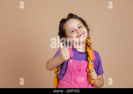 Portrait eines lächelnden kleinen Mädchens mit gelben Kanekalon-Zöpfen mit fehlendem Zahn, das Haar mit der Hand berührt und wie ein Schild mit pinkem Jumpsuit zeigt Stockfoto