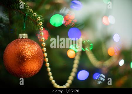 An Weihnachten hängt eine goldorange Weihnachtsbaumkugel an einem Weihnachtsbaum. Glänzende Weihnachtsbaumlichter im Hintergrund. Stockfoto