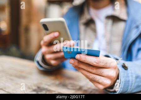 Weicher Fokus von Crop-Mann mit Handy mit Kreditkarte für den Kauf zu bezahlen, während am Tisch sitzen und online einkaufen im Restaurant Stockfoto