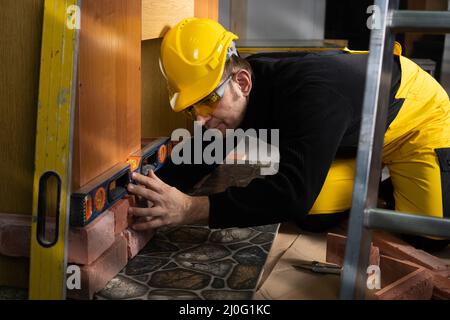 Der Handarbeiter überprüft mit einer Wasserwaage, ob der dekorative Ziegel richtig positioniert ist. Ein Bauarbeiter in der Kleidung Stockfoto