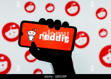 Smartphone mit dem Logo der Reddit-Diskussionsplattform auf dem Bildschirm in einer zusammengeballten Hand auf dem Hintergrund der Reddit-Logos Stockfoto