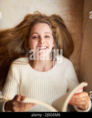 Lesen ist mein Lieblingshobby. Porträt einer attraktiven jungen Frau, die ein Buch liest, während sie zu Hause auf dem Sofa liegt. Stockfoto
